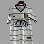 A white Los Angeles Galaxy Nike soccer jersey with black horizontal stripes, featuring the Galaxy crest on the chest, a black Nike swoosh, and a 'GALAXY' sponsor logo across the front, displayed on a wooden hanger against a solid gray background.