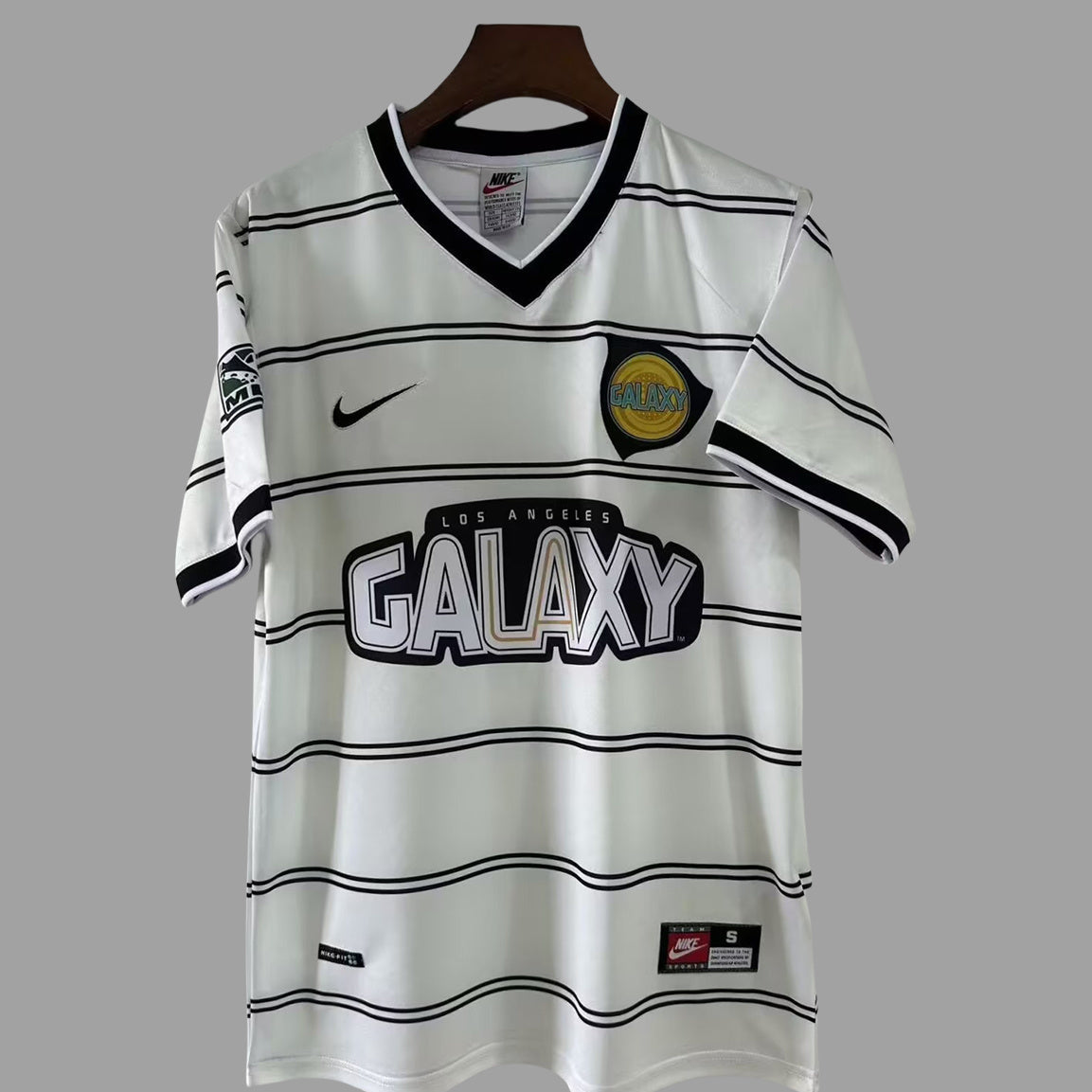A white Los Angeles Galaxy Nike soccer jersey with black horizontal stripes, featuring the Galaxy crest on the chest, a black Nike swoosh, and a 'GALAXY' sponsor logo across the front, displayed on a wooden hanger against a solid gray background.