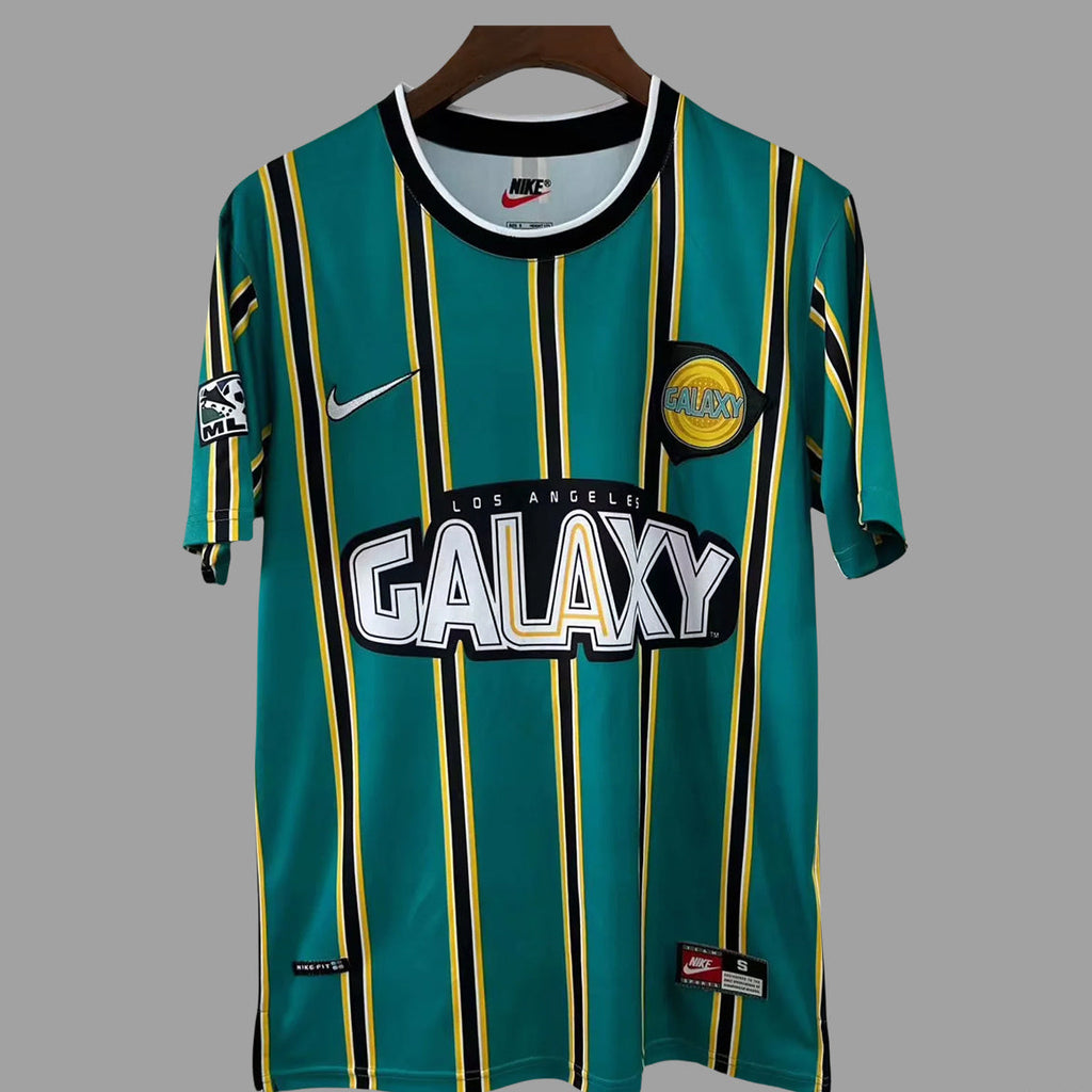 A Nike Los Angeles Galaxy soccer jersey in teal green with vertical yellow and black stripes, featuring the 'Los Angeles Galaxy' team crest and 'GALAXY' sponsor logo on the front, hanging on a wooden hanger against a solid gray background.