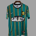 A Nike Los Angeles Galaxy soccer jersey in teal green with vertical yellow and black stripes, featuring the 'Los Angeles Galaxy' team crest and 'GALAXY' sponsor logo on the front, hanging on a wooden hanger against a solid gray background.