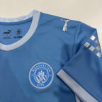 Light blue Manchester City home jersey with crest, Puma branding, and checkered sleeve patch