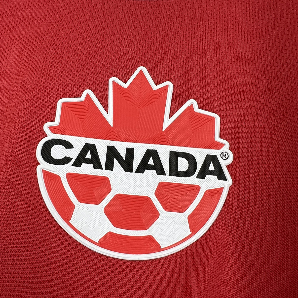 Red Canada crest badge on soccer jersey with white outline and red maple leaf.