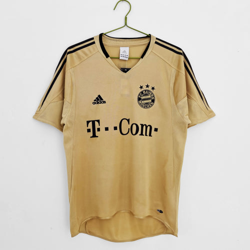 Gold football jersey on hanger with V-neck, adidas mark, Bayern crest, and T‑Com sponsor.